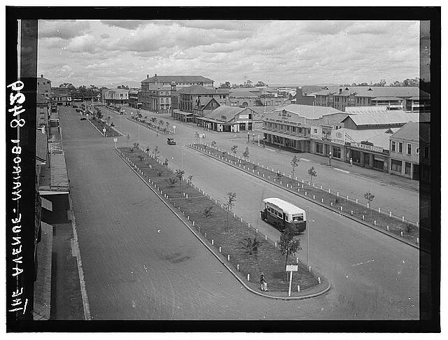 One, two, and three story commercial buildings flank a street. There are medians planted with trees divide the roadway into three sections. A bus and a car are present on the street. There are sidewalks and curbs along what appears to be a dirt surfaced roadway.