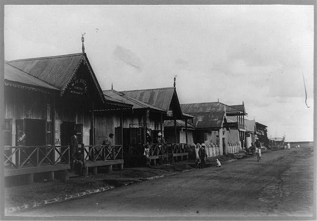 One story frame houses with wooden front porches line a dirt road with stone gutter. Planks provide access from roadway across the gutter to wooden front porches. People stand on the porches, a man rides a bicycle in the street, a dot sits in the street.
