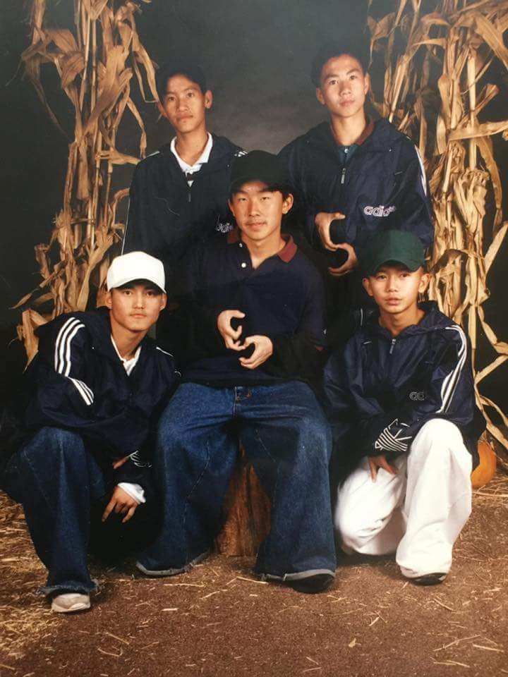 Five men pose in front of a backdrop of browned cornstalks. They all wear matching navy adidas jackets, three wear baseball caps. Two pose with hands held in front of them making what looks like an "S"