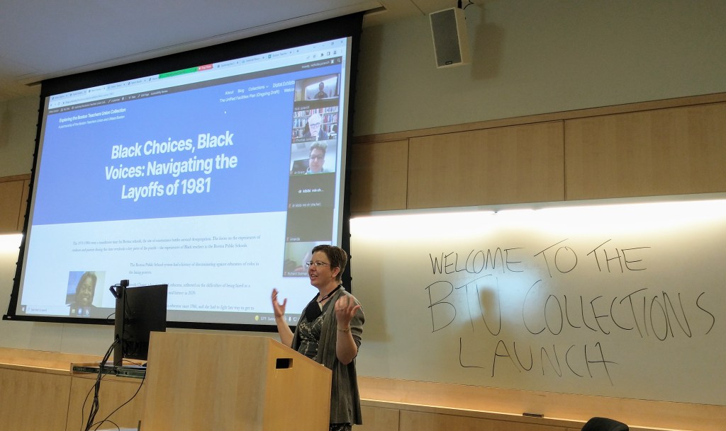 A woman stands at a podium. On a projector screen behind her to the left are the words "Black Choices, Black Voices: Navigating the Layoffs of 1981" and images of people on a video call. On a whiteboard behind her to the right is written "Welcome to the BTU Collections Launch" in large lettering.
