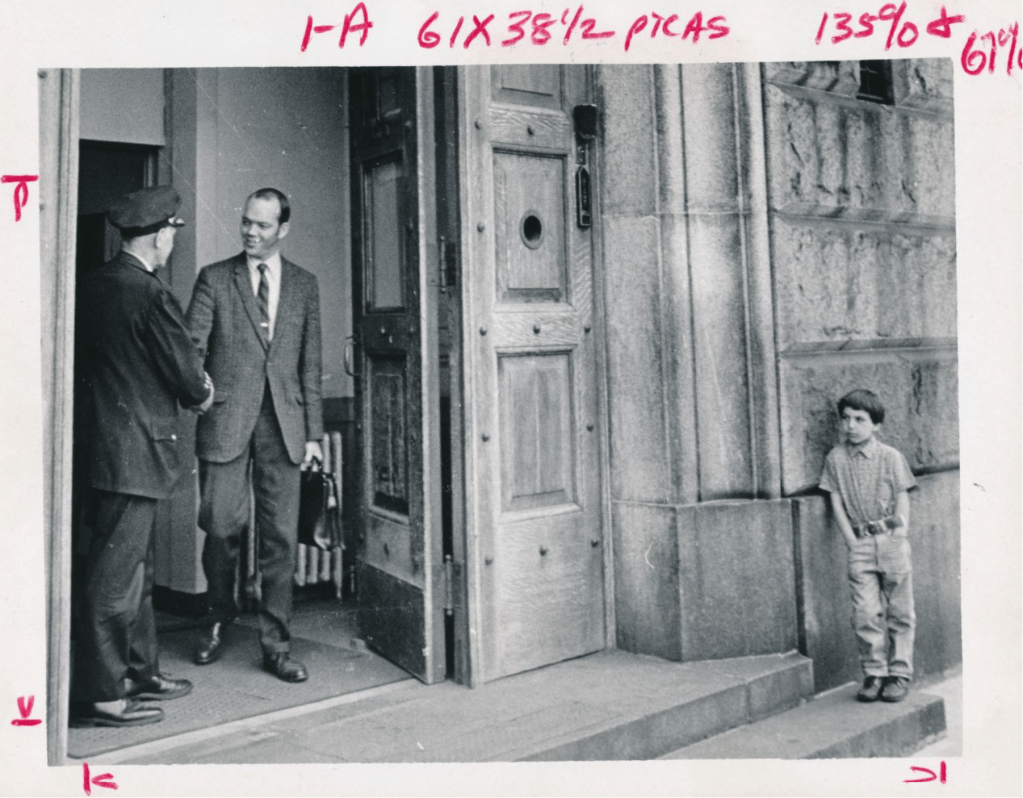 Black and white photo of a  white man  in a suit and tie, carrying a briefcase, walking out of a building and shaking the hand of a white man in uniform with hat. Outside the building, just beside the door, a white boy leans his shoulder against the building, one step down from the door and one step up from the sidewalk, facing toward the door, hands in trouser pockets. The door is very large and made of wood. The building is constructed of very large blocks of stone.