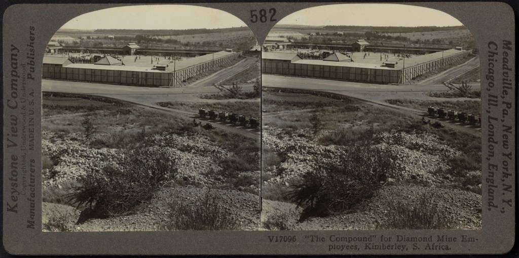 A black-and-white stereogram shows two identical side-by-side images with shrubland and a rail line with empty open-topped railcars in the foreground and a rectangular enclosure in the background. The enclosure has thick walls and people milling about in the open space inside them. The stereograph is labeled “‘The Compound’ for Diamond Mine Employees, Kimberley, S. Africa” on the bottom and “Keystone View Company” on the left side.