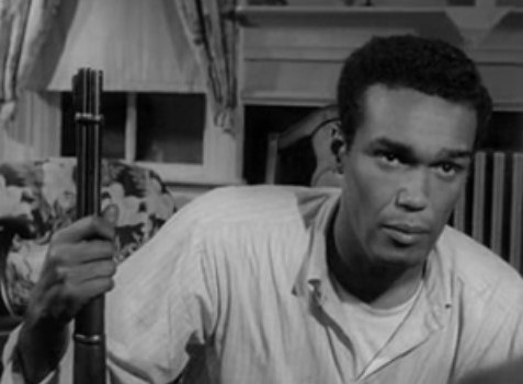 Black and white photograph of African American actor Duane Jones, leaning to his left while  holding a gun in his right hand in what appears to be the living room of a house.