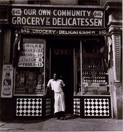 Black and white photograph from 1940 of an Arican American man wearing apron standing in doorway of Harlem grocery store, with sign, "Our Own Community Grocery & Delicatessen," above.